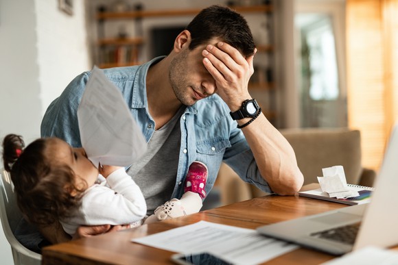 Man at home office with his hand over his forehead while clutching his daughter in his other arm.