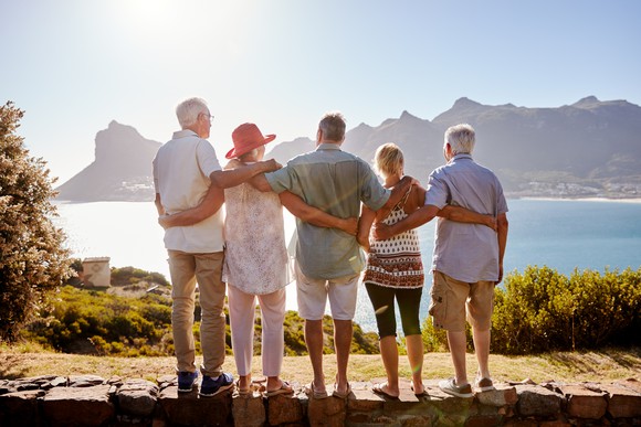 A group of retirees with arms around each other on the top of a cliff overlooking a bay and mountains viewed from behind. 