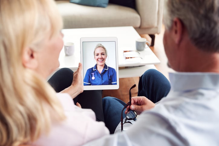 Mature male and female sitting in a house and holding a computer tablet with a young female nurse on the screen.