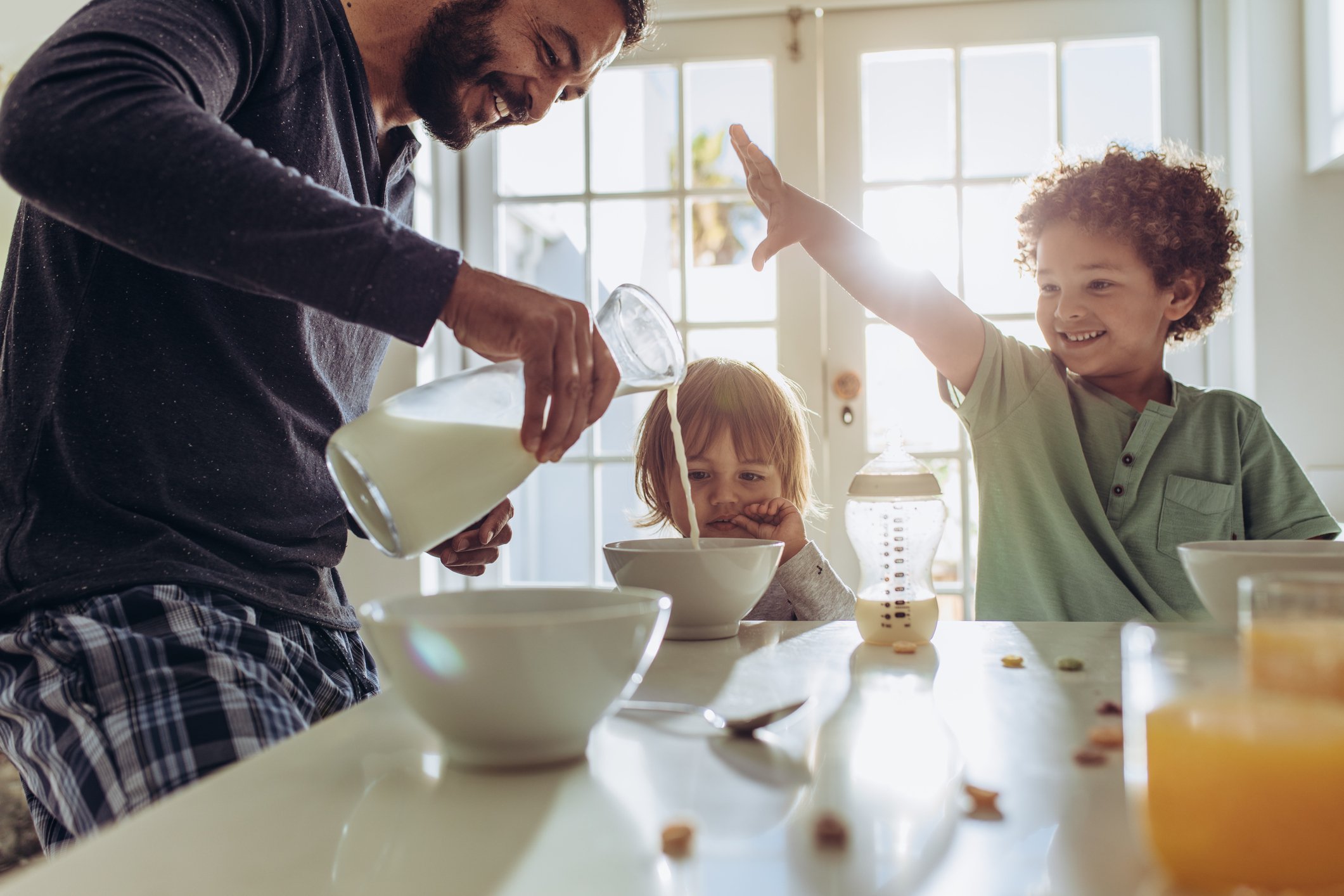 Man pouring milk into cereal for two kids at a table.