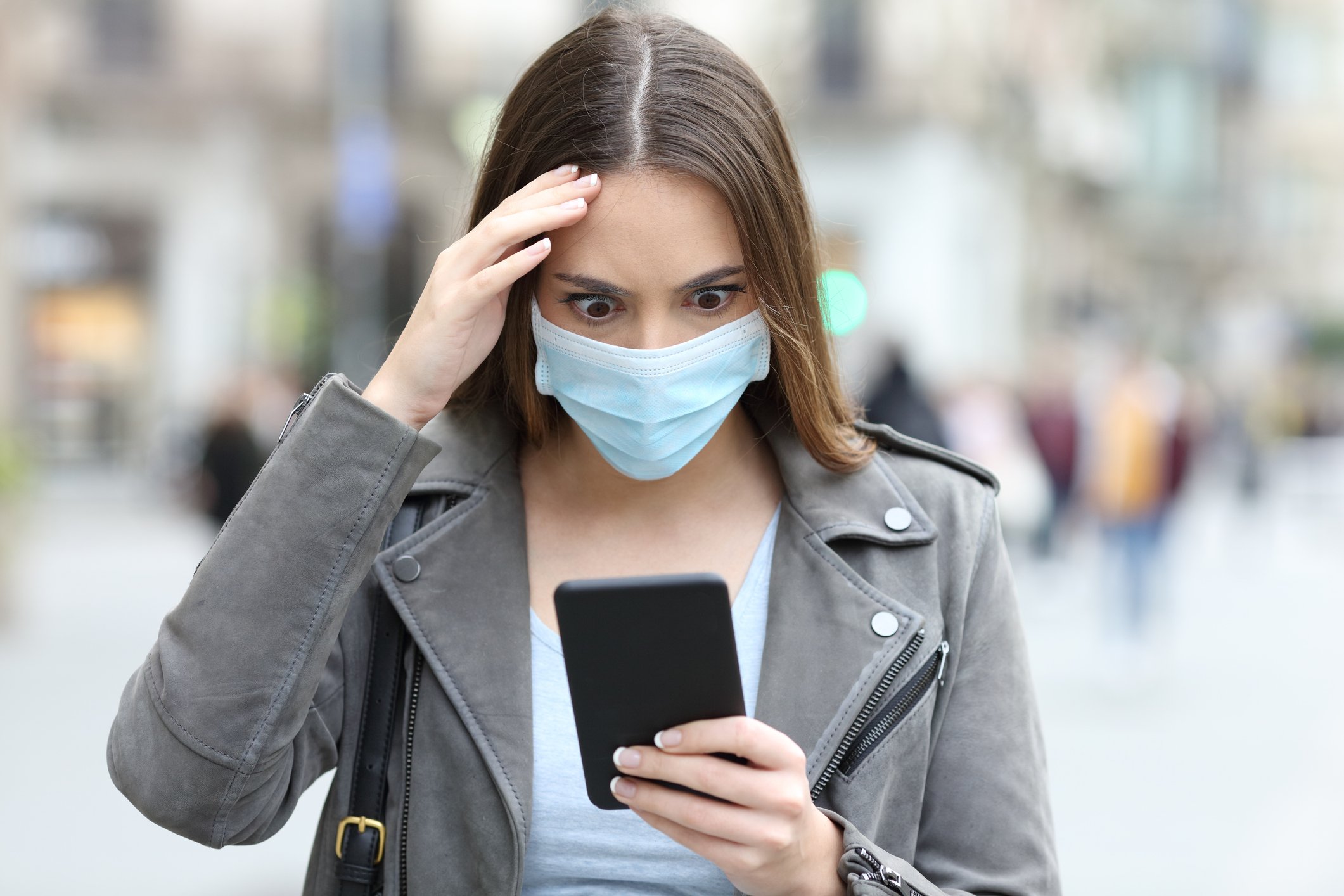 A young woman, wearing a medical face mask, stares wide-eyed at her phone.