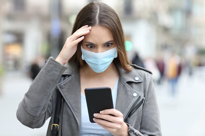 A young woman, wearing a medical face mask, stares wide-eyed at her phone.