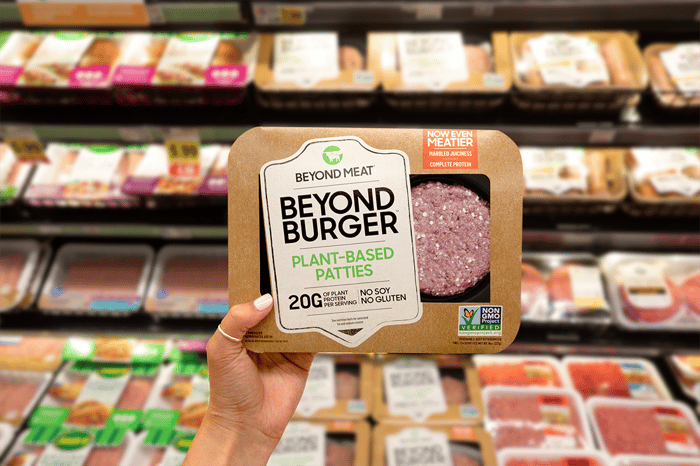 Hand holding up a Beyond Meat patty package in front of a grocery store meat case