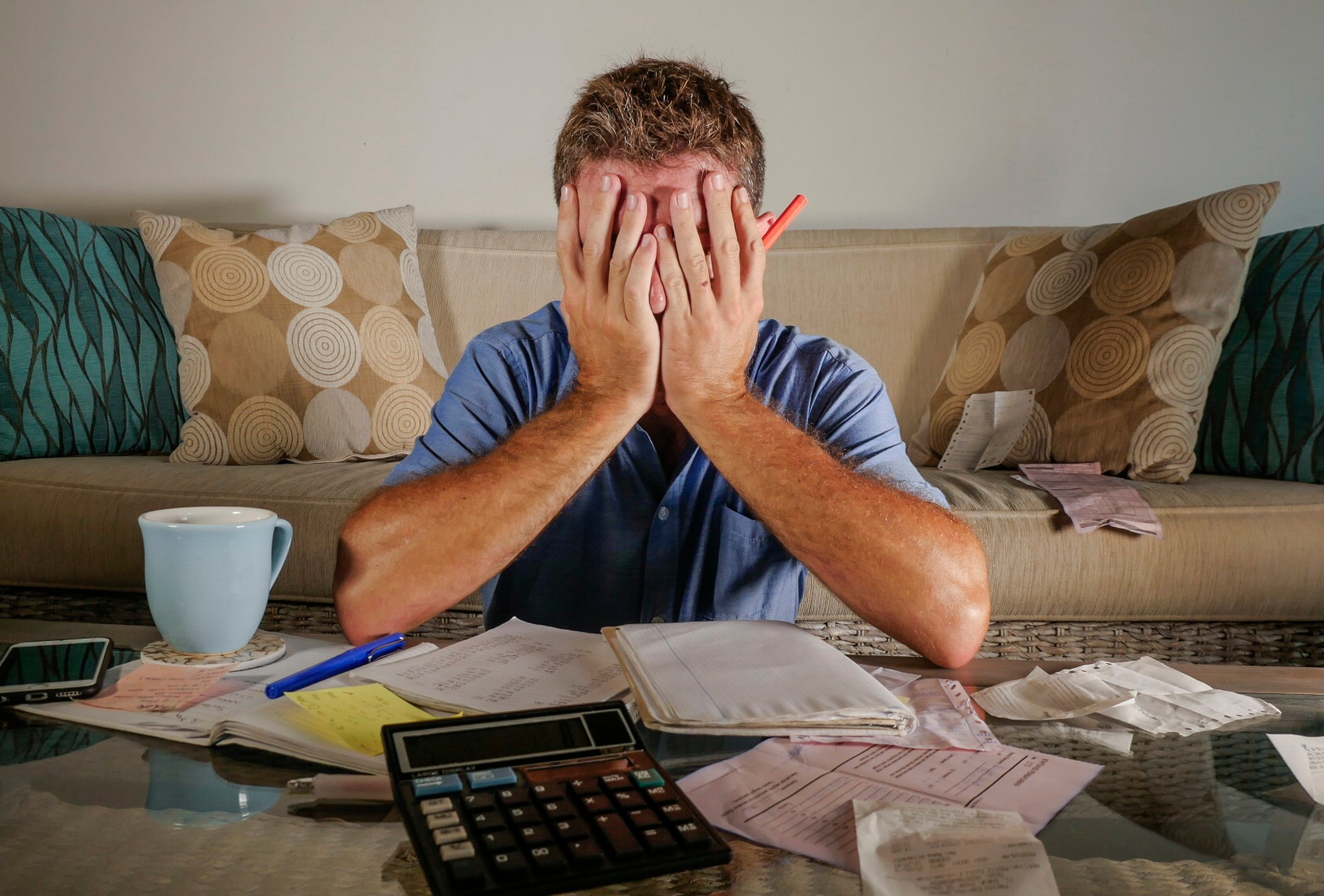 Man covering his face; spread out on a table in front of him are documents, a calculator, a pen, and a mug