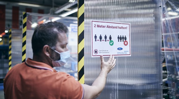 A worker wearing a mask is shown next to a sign reminding employees to maintain social distancing at a Ford factory in Germany.