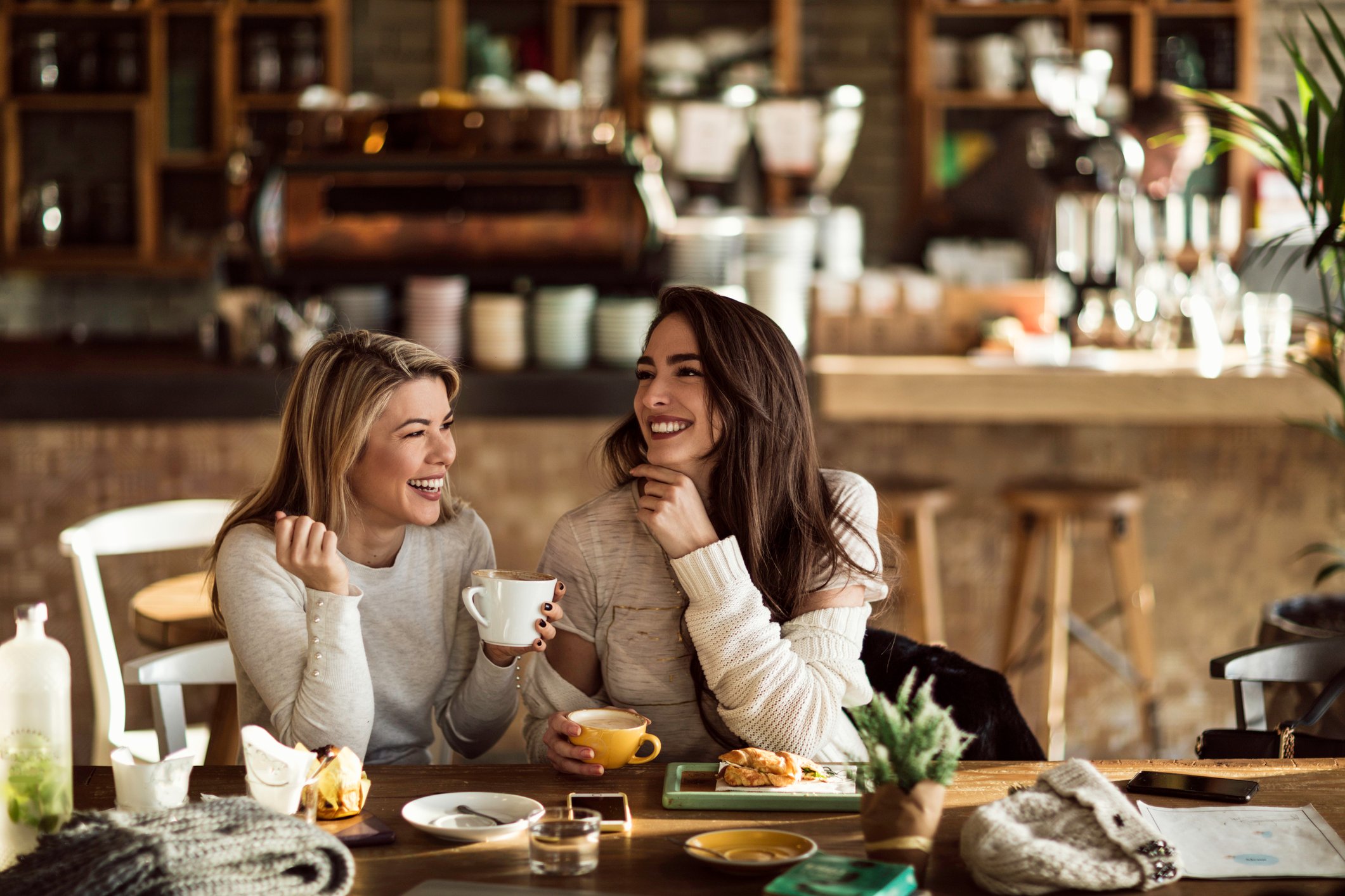 Two women smiling in a coffee shop while holding their mugs.