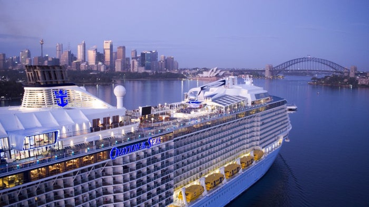 Cruise ship in a port with a city skyline and rainbow bridge nearby.