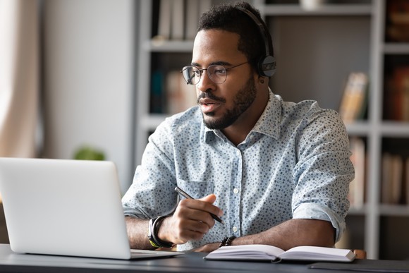 A man wearing headphones and looking at his laptop screen while also holding a pen near a notebook