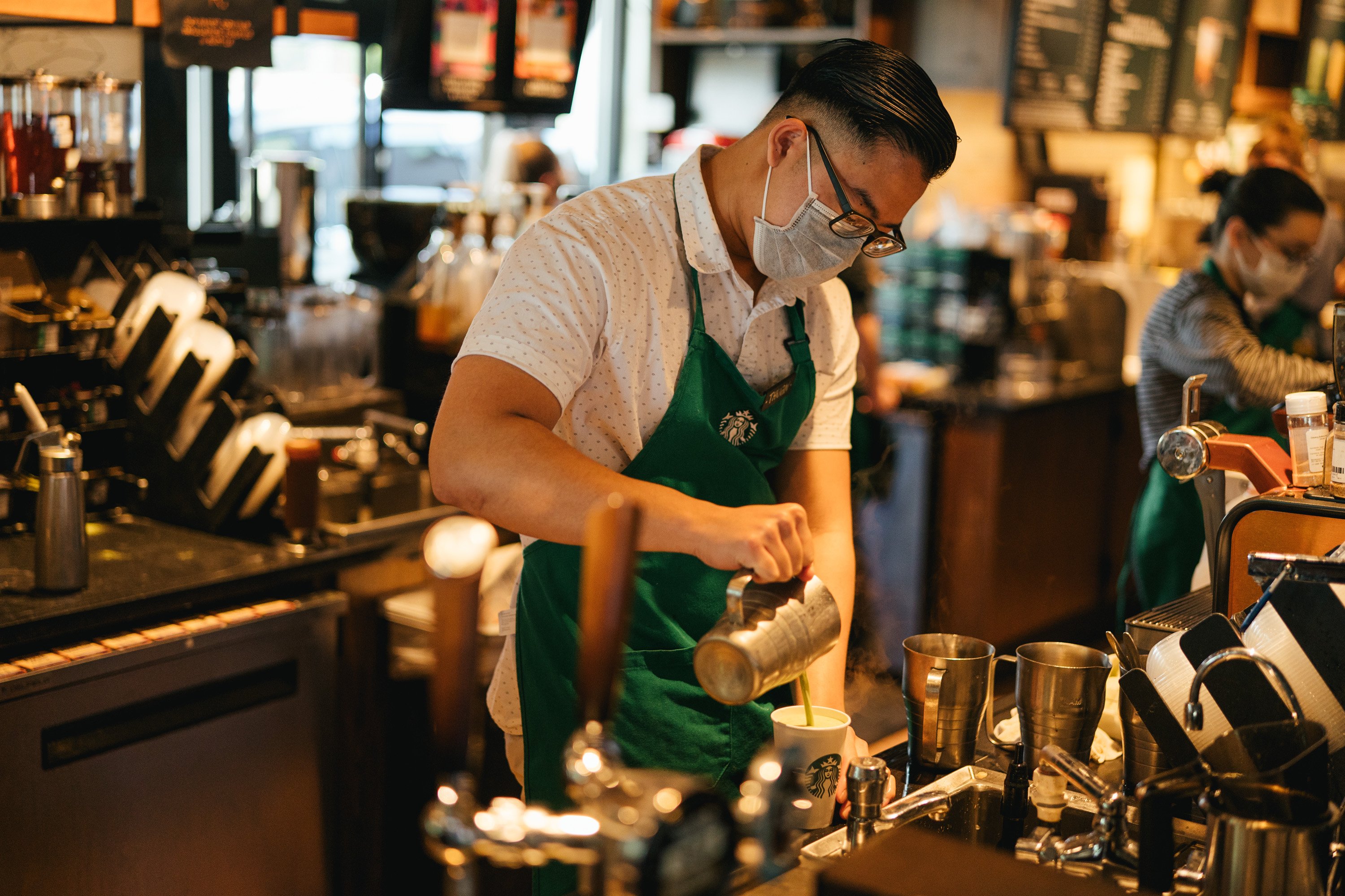 A Starbucks employee, wearing a mask, prepares a beverage at the counter.