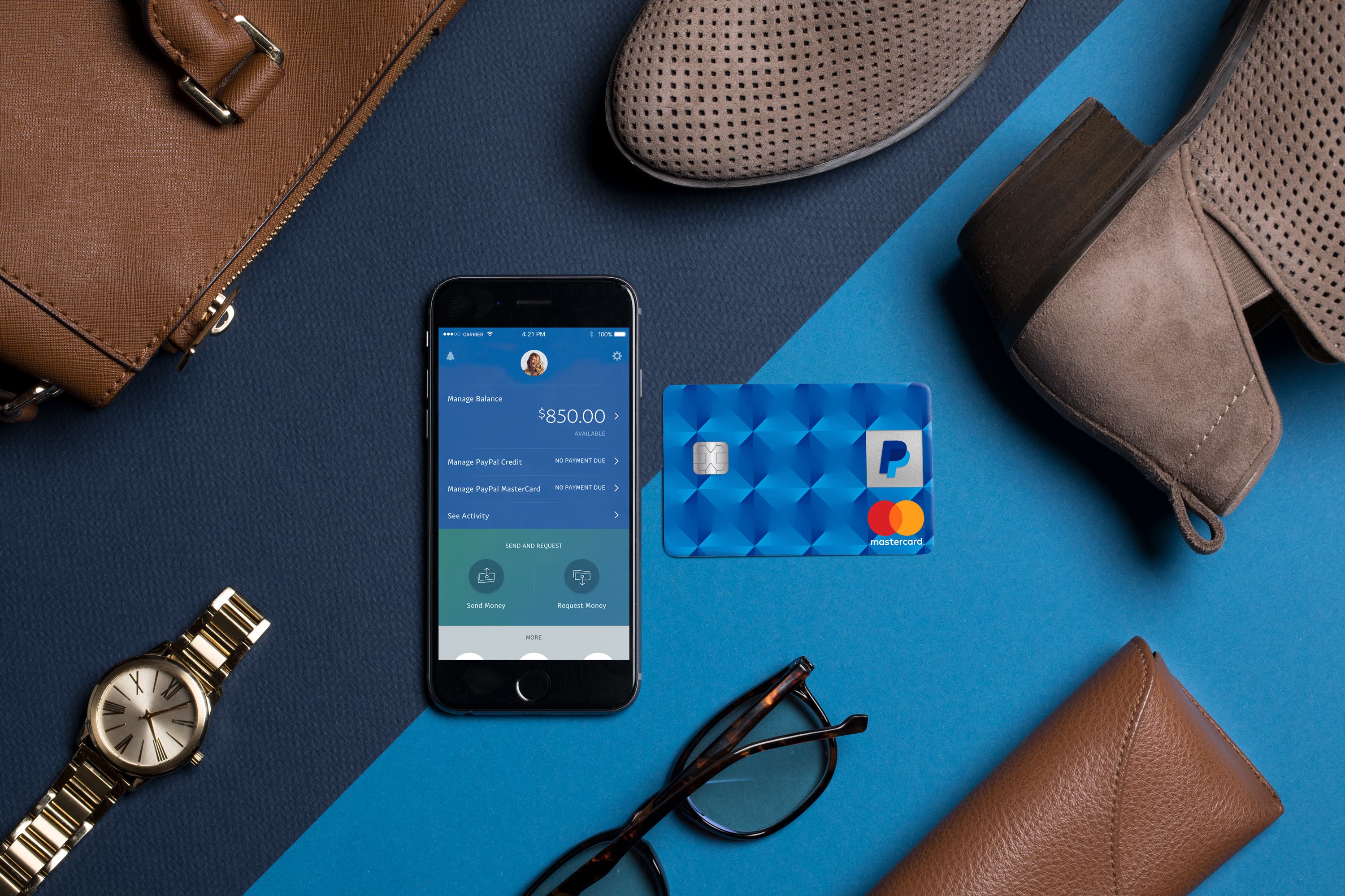 A smartphone displaying the PayPal app is surrounded by items such as a credit card, shoes, and a watch.