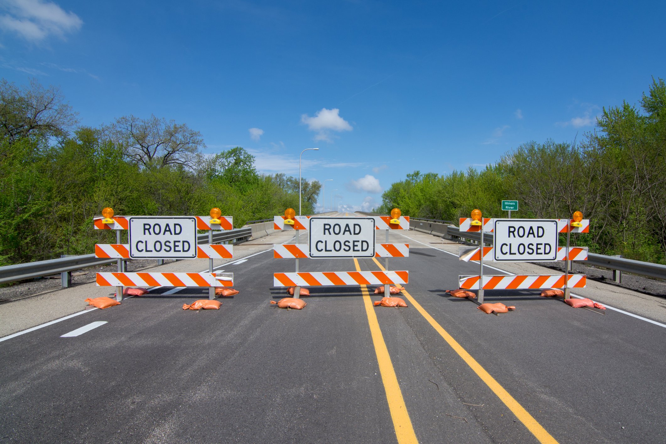 three roadblock signs sit on an open road with woods and blue sky in the background.