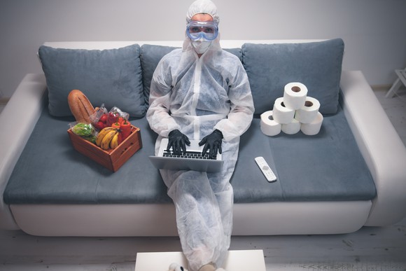 A woman in protective gear sitting on the couch with her laptop with toilet paper and produce beside her.