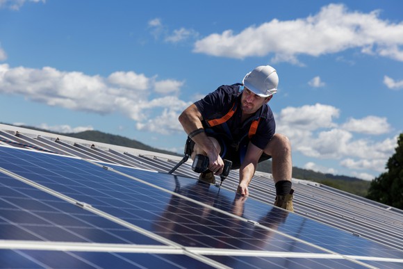 A solar panel installer works with a drill on a roof.