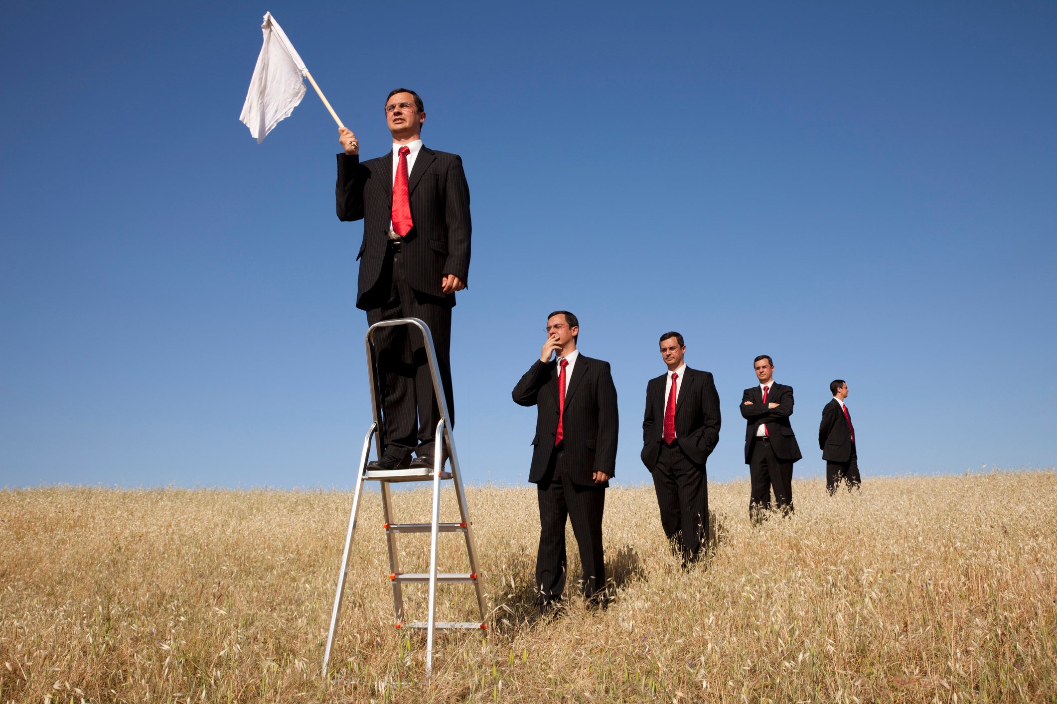 A group of businessmen in a field, getting in line to stand on a stepladder and wave a white flag.