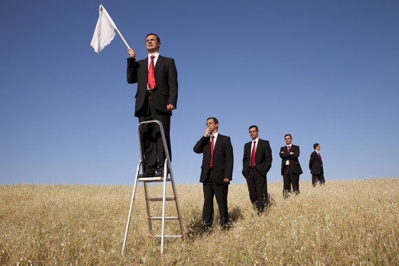 A group of businessmen in a field, getting in line to stand on a stepladder and wave a white flag.