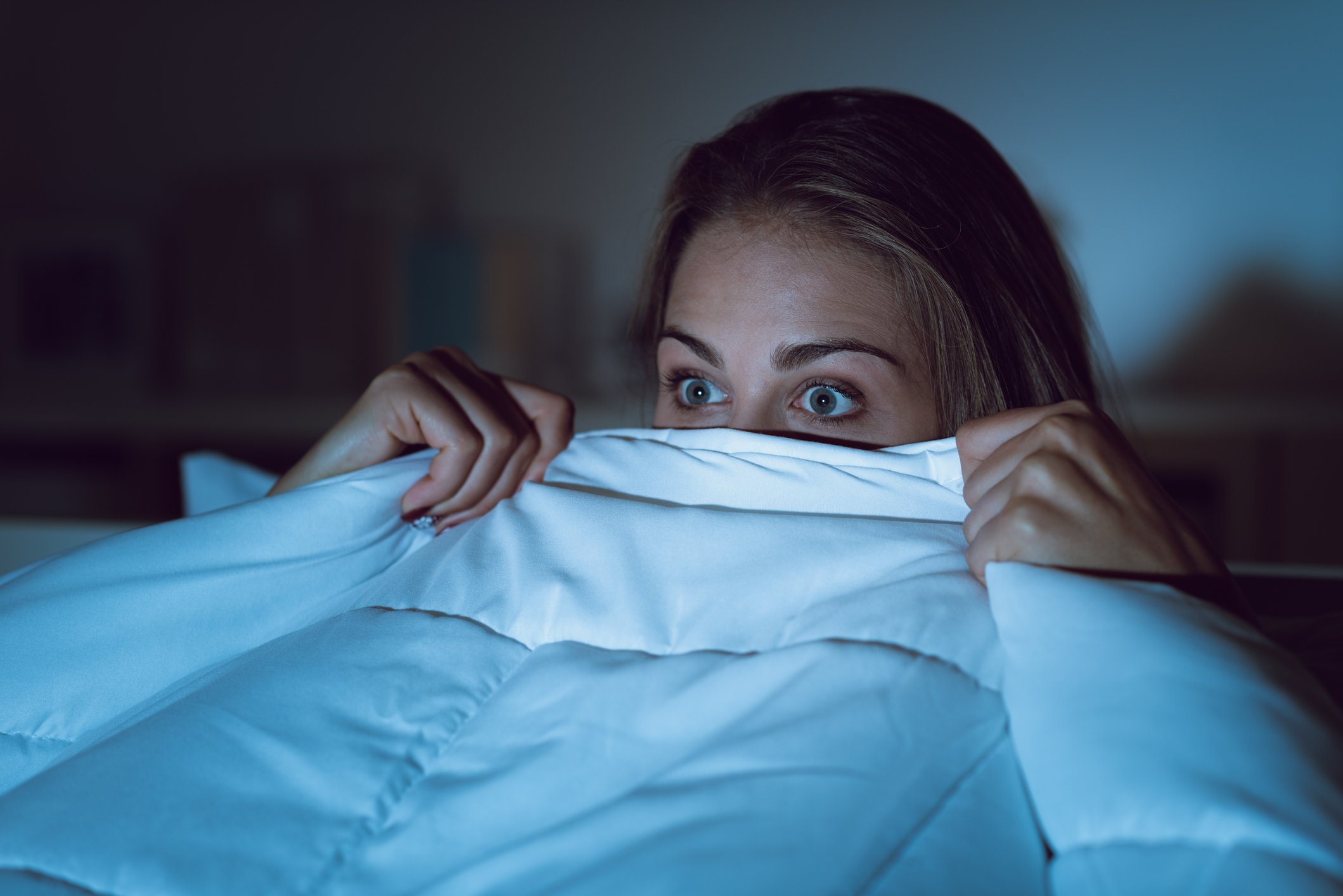 Scared woman peeking out from under bed covers.