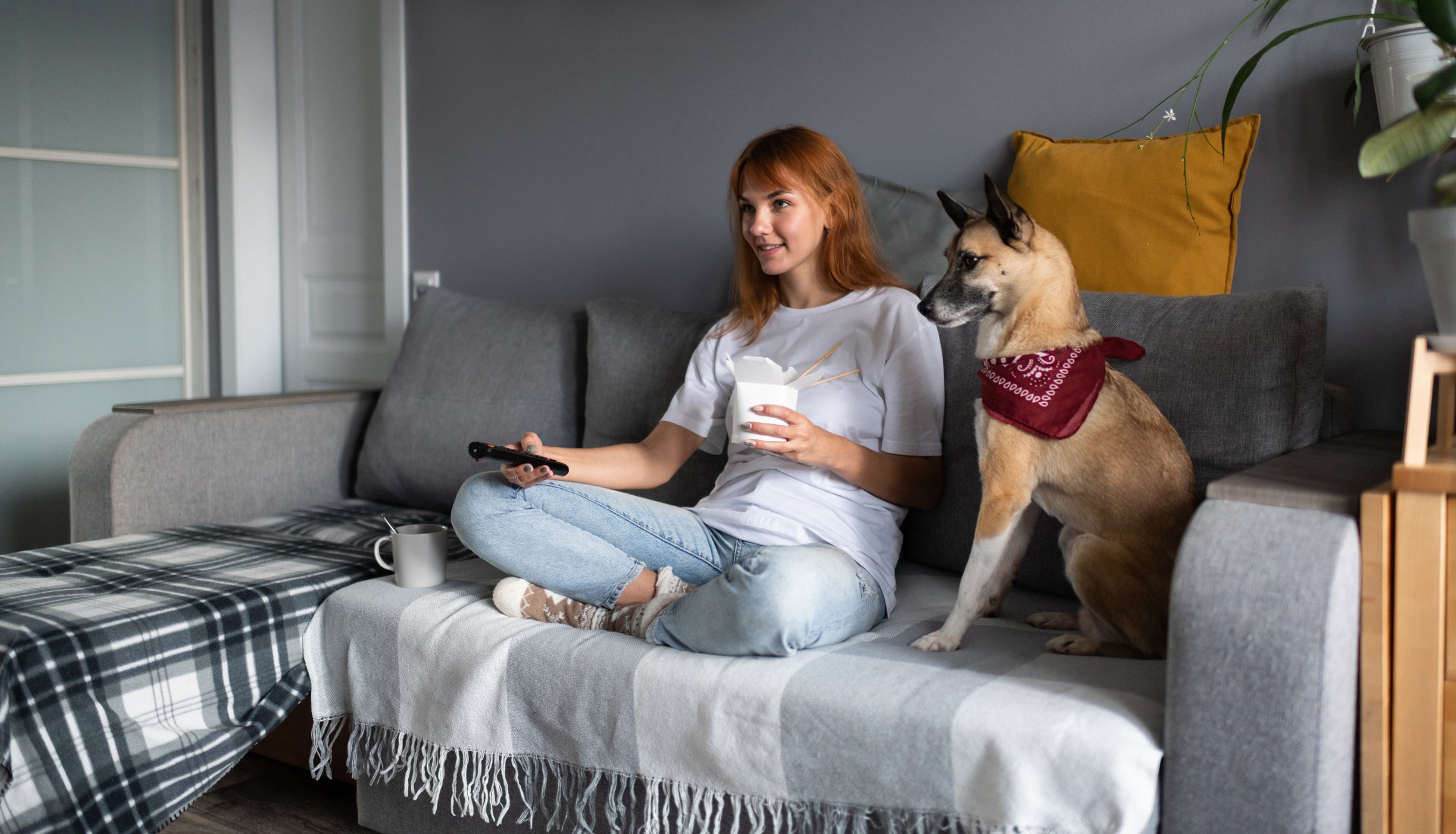 Woman sitting on couch next to a dog while holding a TV remote