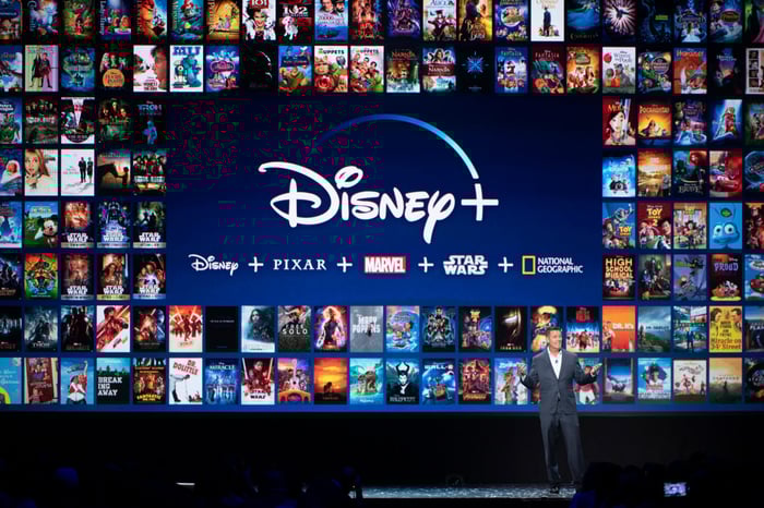 A man standing in front of a presentation screen featuring dozens of title cards for Disney+.