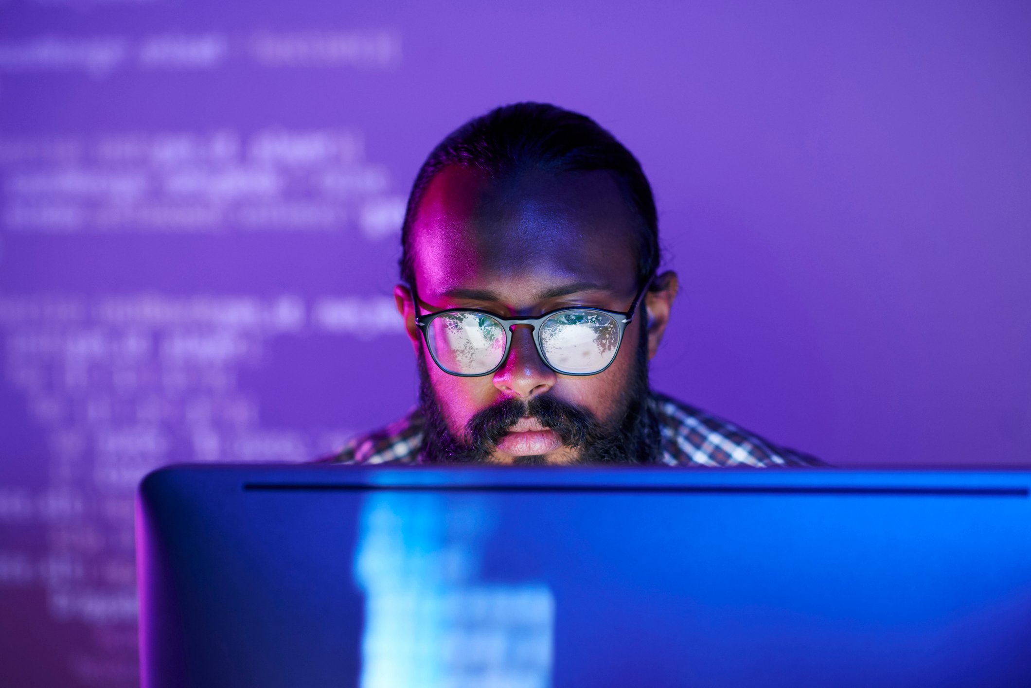 Young man in glasses looks  at a desktop screen in  a dark room lit with purple light. 