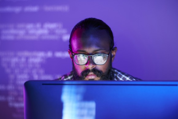 Young man in glasses looks  at a desktop screen in  a dark room lit with purple light. 