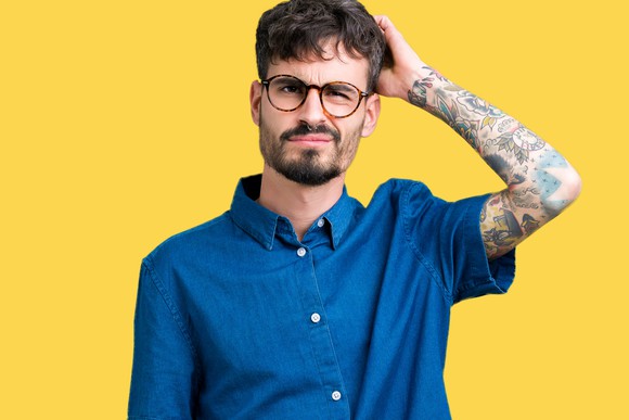 A young hipster with tattoos and glasses scratches his head in confusion. 