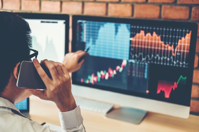 A man on his cell phone pointing at a computer screen filled with stock market data