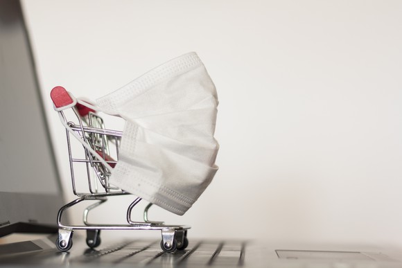 A mini shopping cart with a face mask on it is shown on the keyboard of a laptop. 