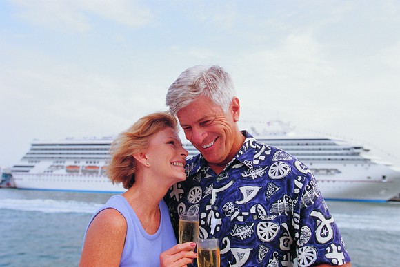 An older man and woman holding glasses of champagne with a cruise ship in the background