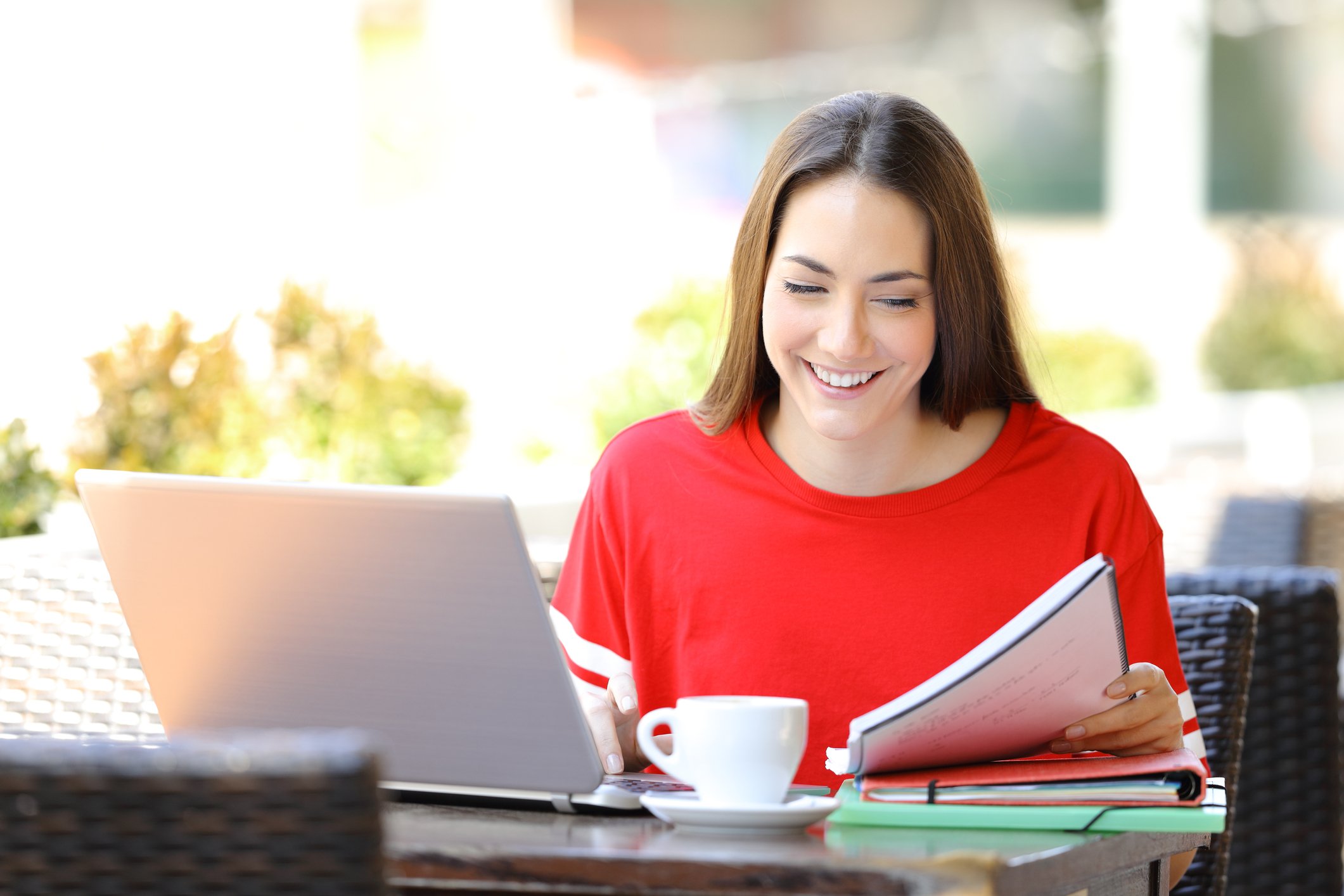 A young woman in a red shirt smiles at her laptop and notebook.