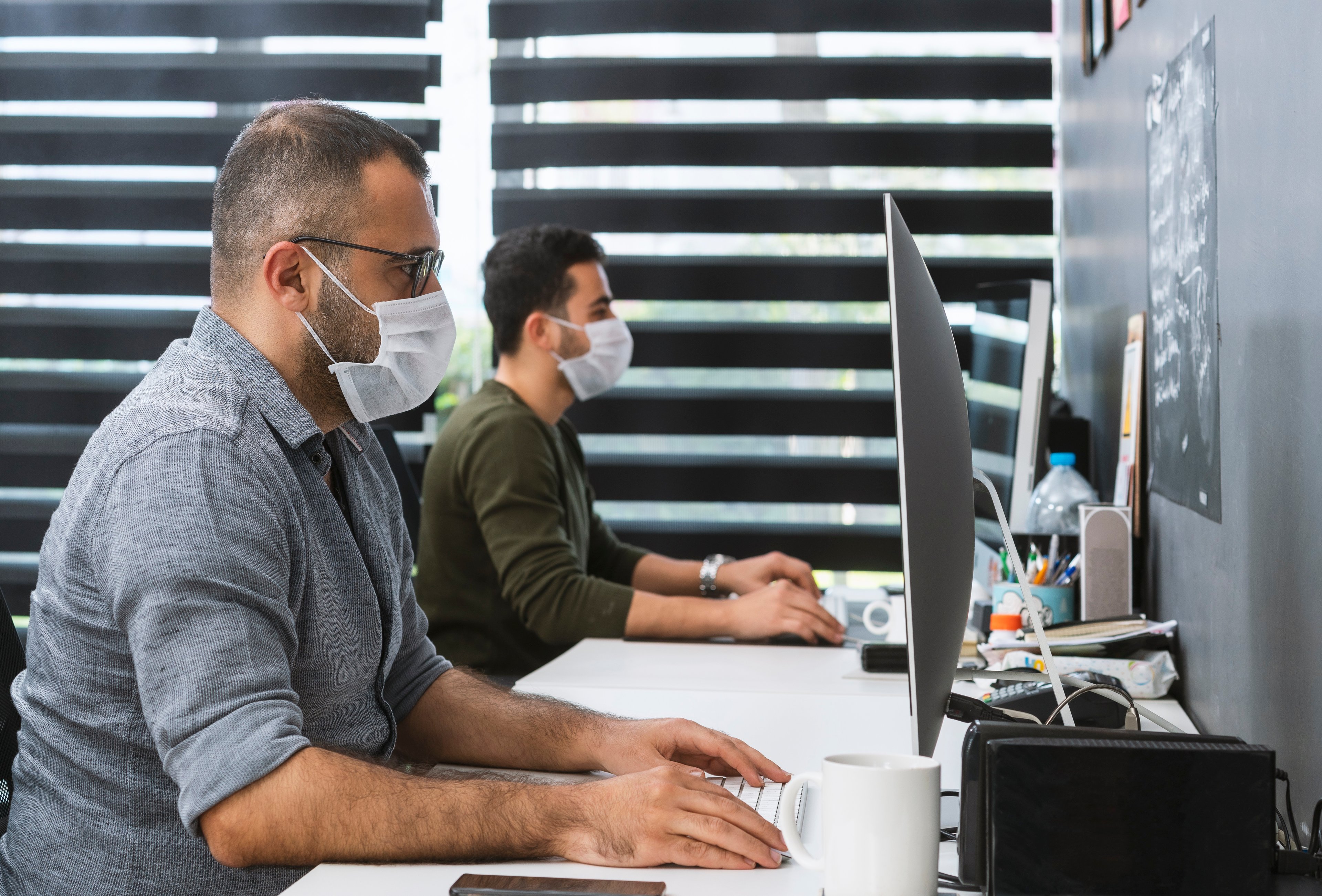 Two men working at computers in an office wearing surgical masks. 