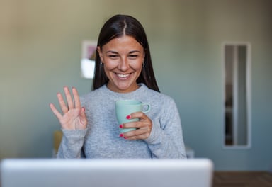 Smiling woman holding mug waves at laptop screen