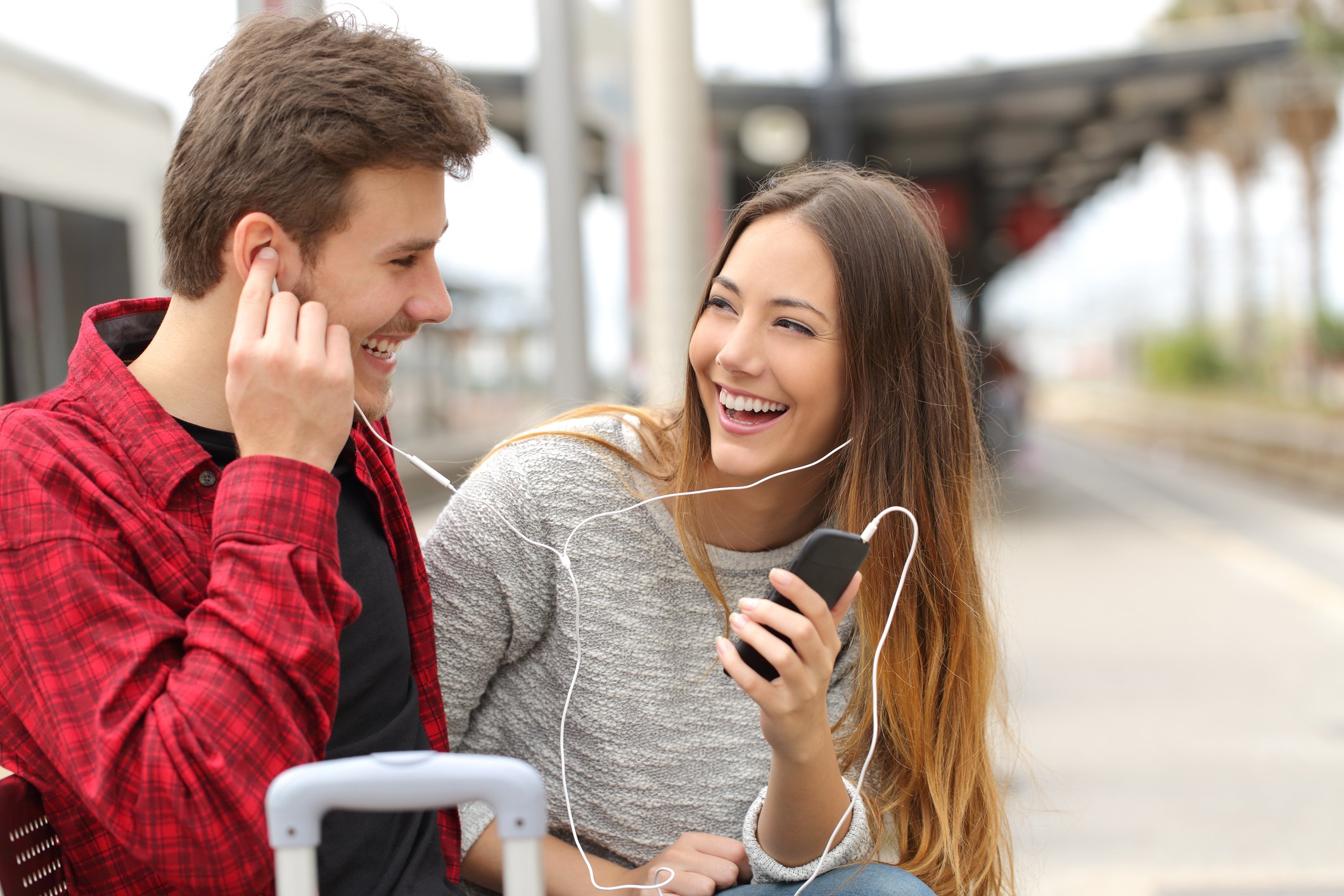 A young couple smile at each other, sharing a pair of headphones attached to one of their smartphones.