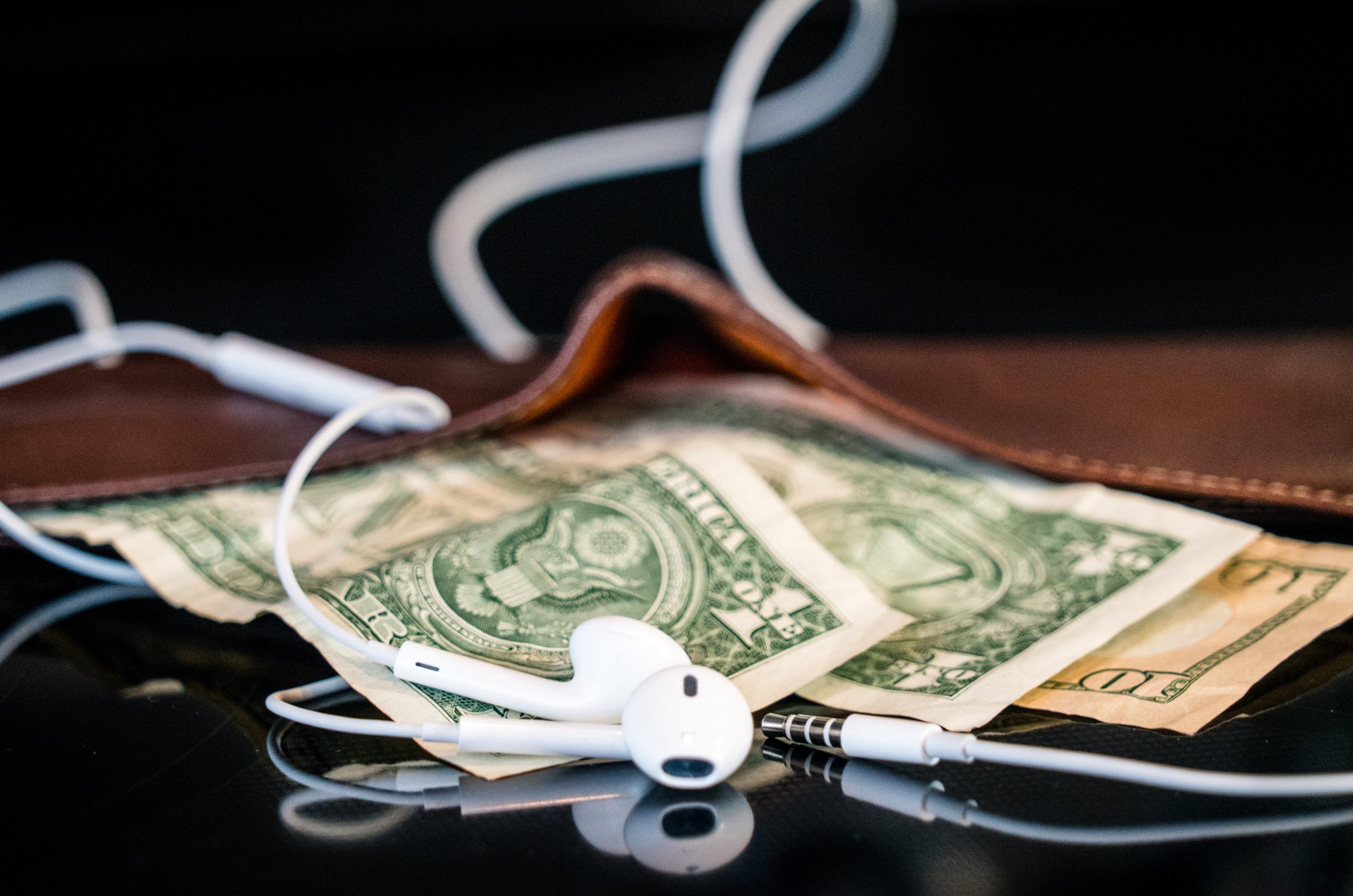 A pair of white earbuds nestled on top of some dollar bils sticking out of a leather wallet.