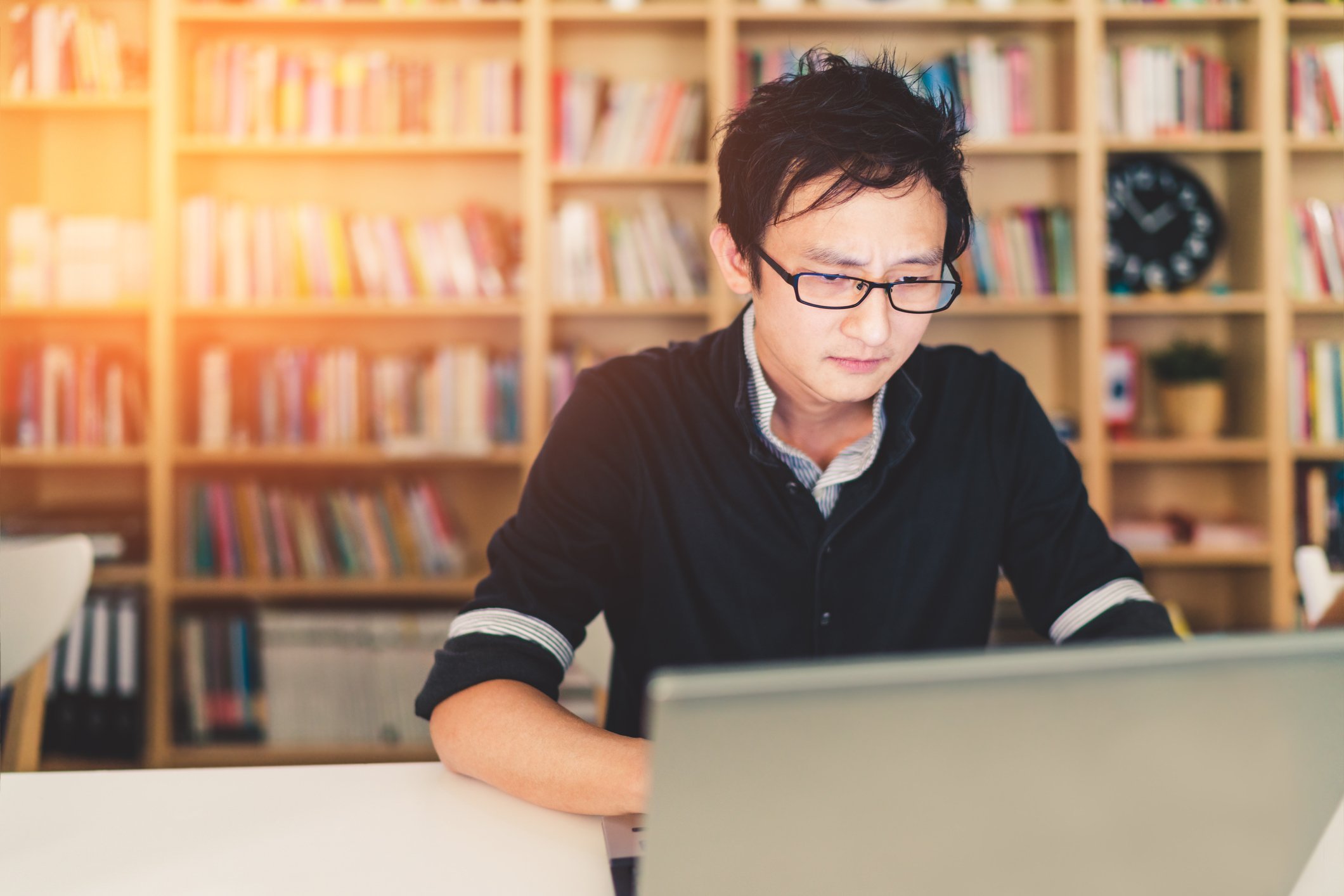 A man on his laptop with a bookcase behind him. 