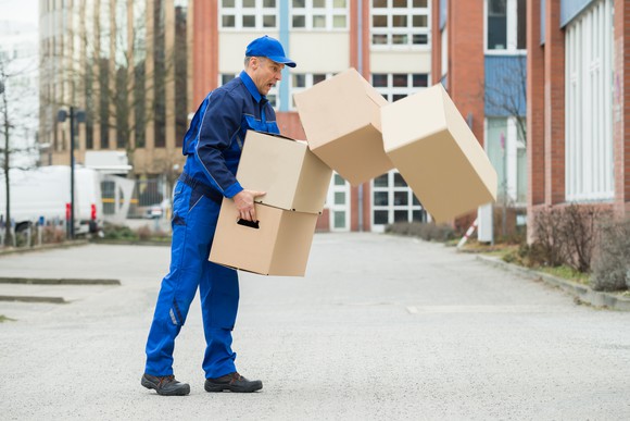 Uniformed delivery man standing on a sidewalk and reacting in surprise as the boxes he's carrying are tumbling out of his grasp.