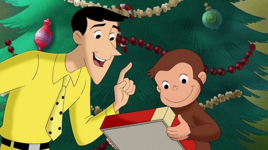 Curious George and character known as The Man with the Yellow Hat stand in front of Christmas tree and ornaments.