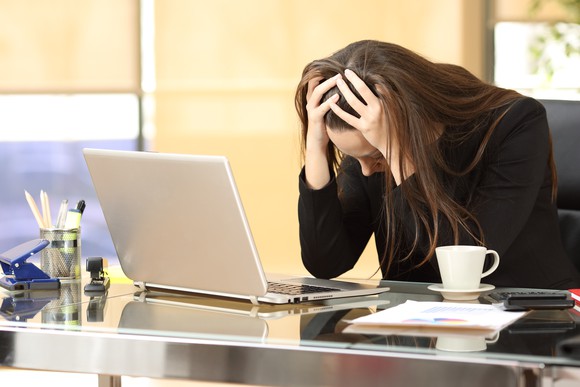 A woman sits at a desk, with her head in her hands and elbows on the desk.