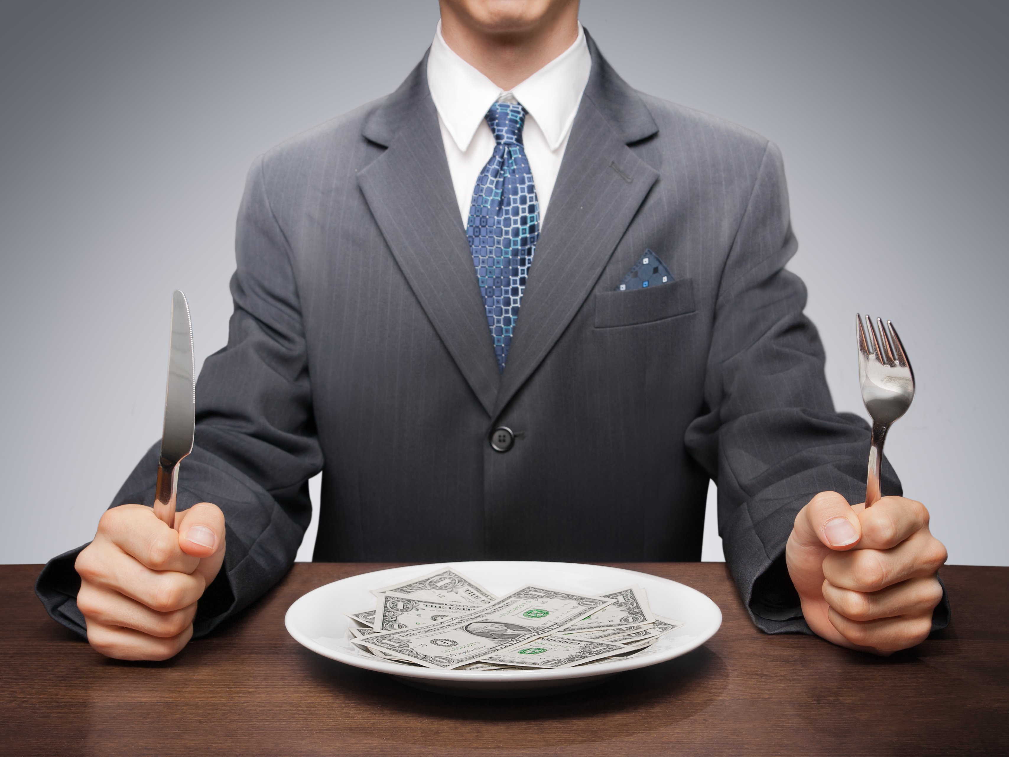 A man in a suit holding a knife and fork sits behind a plate full of $1 bills. 