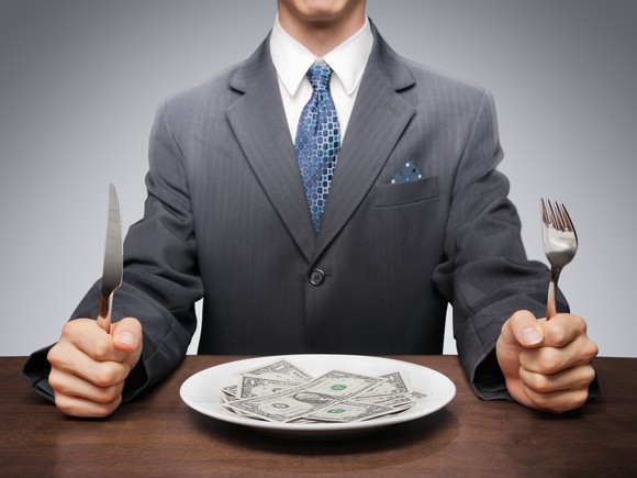 A man in a suit holding a knife and fork sits behind a plate full of $1 bills. 