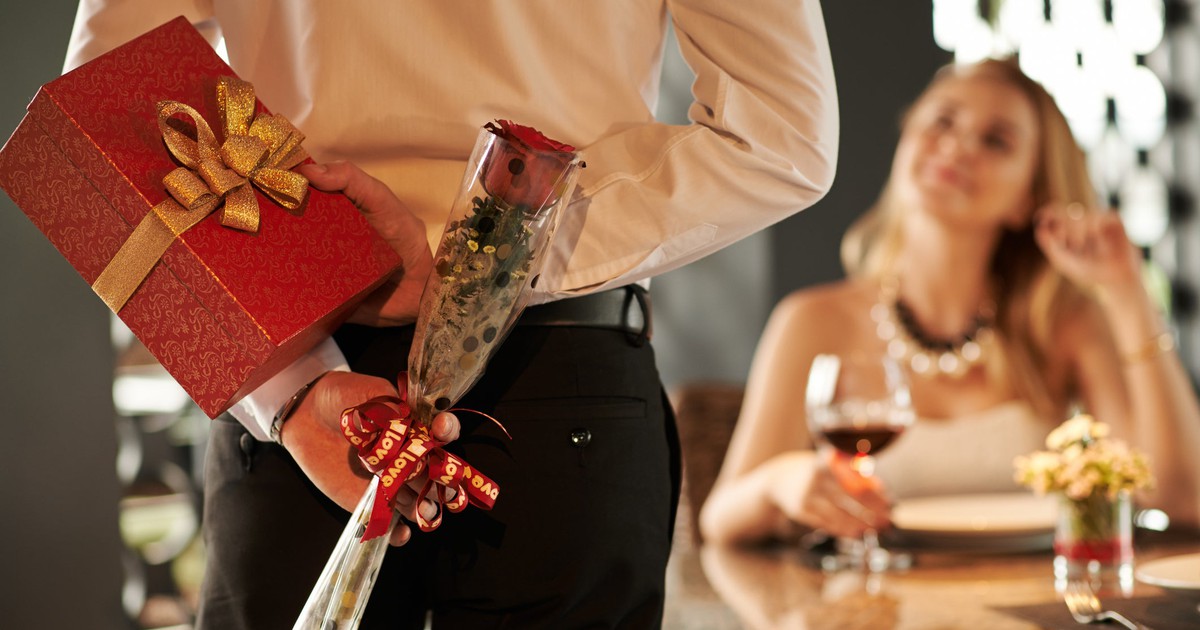 A male date shows up holding a gift box and rose behind his back as he approaches a woman enjoying wine at a table.