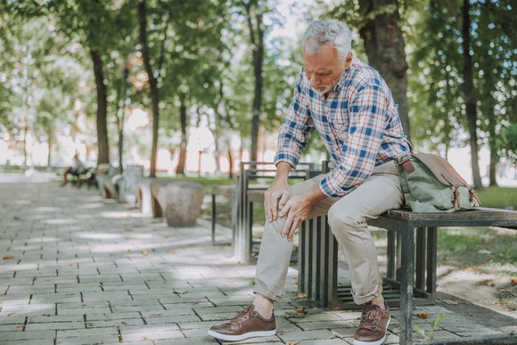 A senior man rubs his knee while sitting on a park bench.
