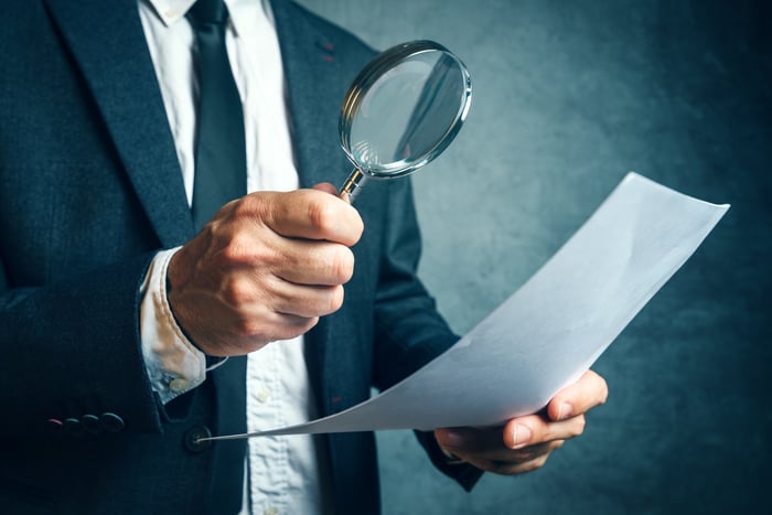 A person in a business suit is inspecting a piece of paper with a magnifying glass.