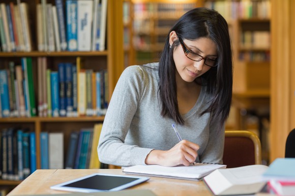 A woman at a desk n a library writes in a notebook.