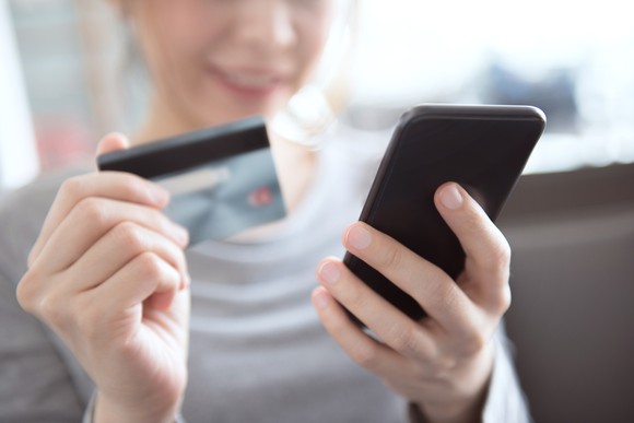 A woman holds up her credit card as she places a shopping order on her phone.
