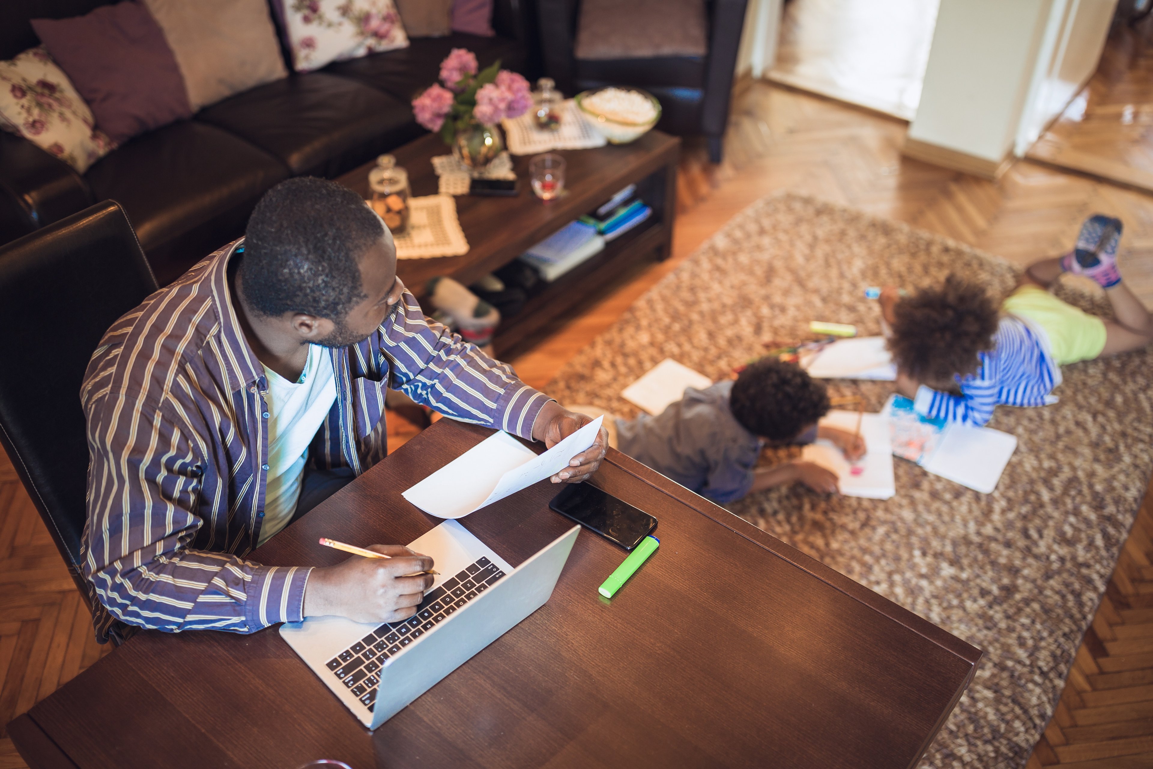 A man sitting at a desk in the living room working with kids coloring on the floor. 
