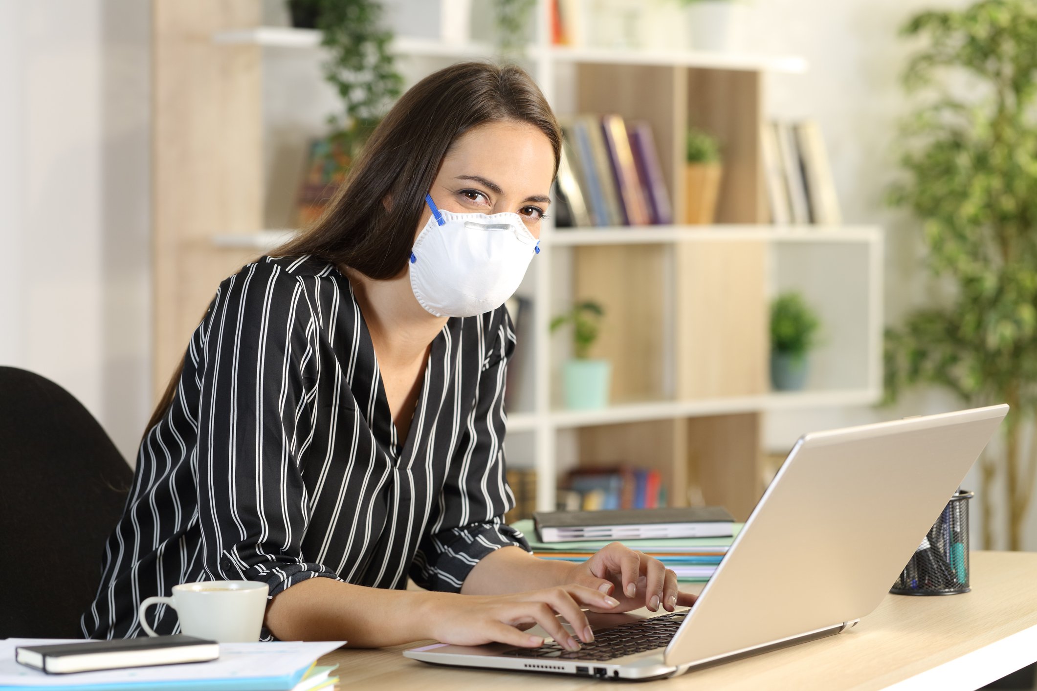 A young woman wearing a face mask works with a laptop at her living room table.