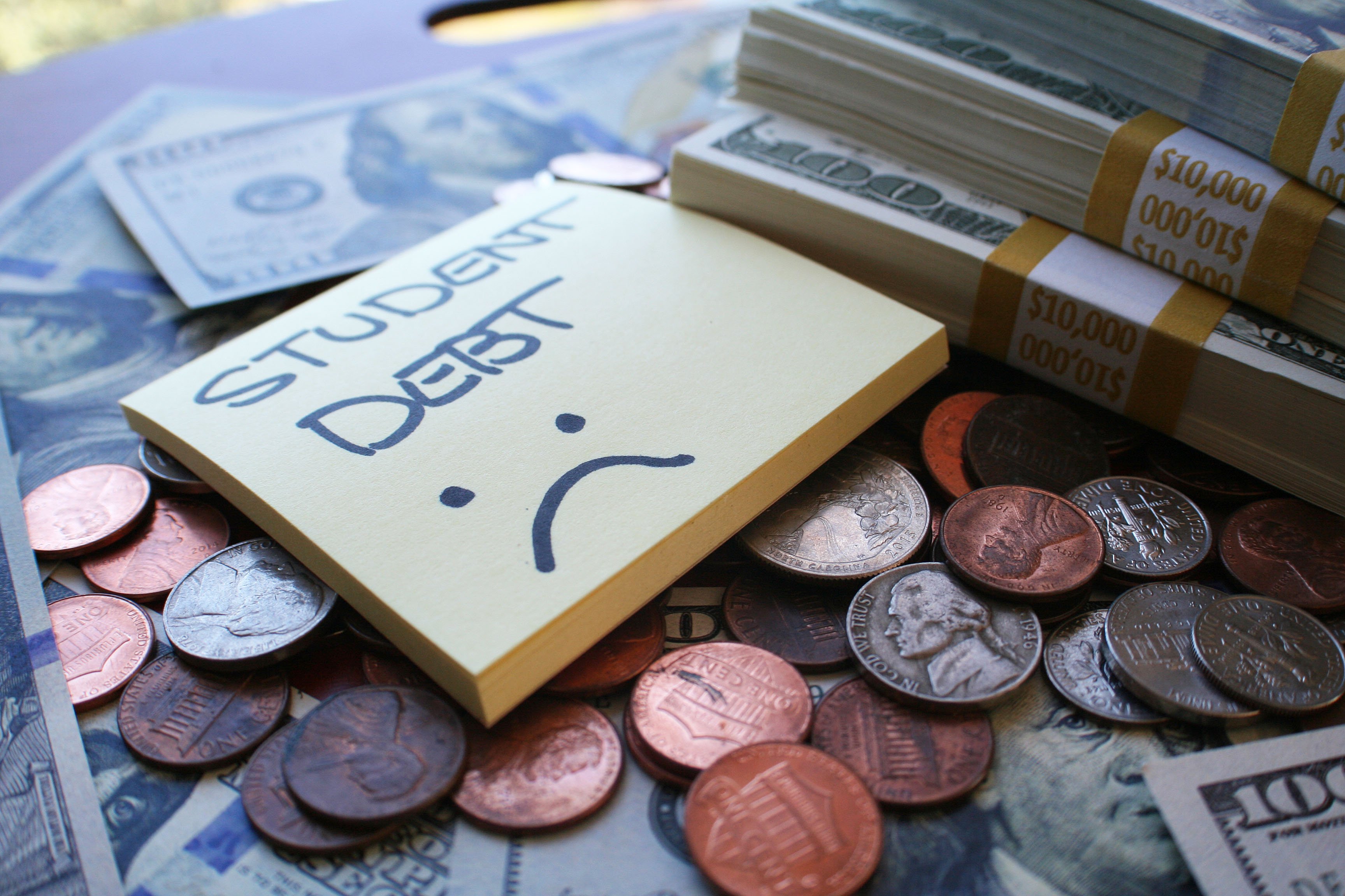A sticky note pad with the words student debt on it over a sad face. The pad sits atop a pile of change and stack of bills.