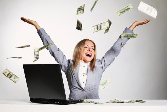 A yong woman smiles and cheers while tossing hundred-dollar bills up in the air in front of an open laptop.