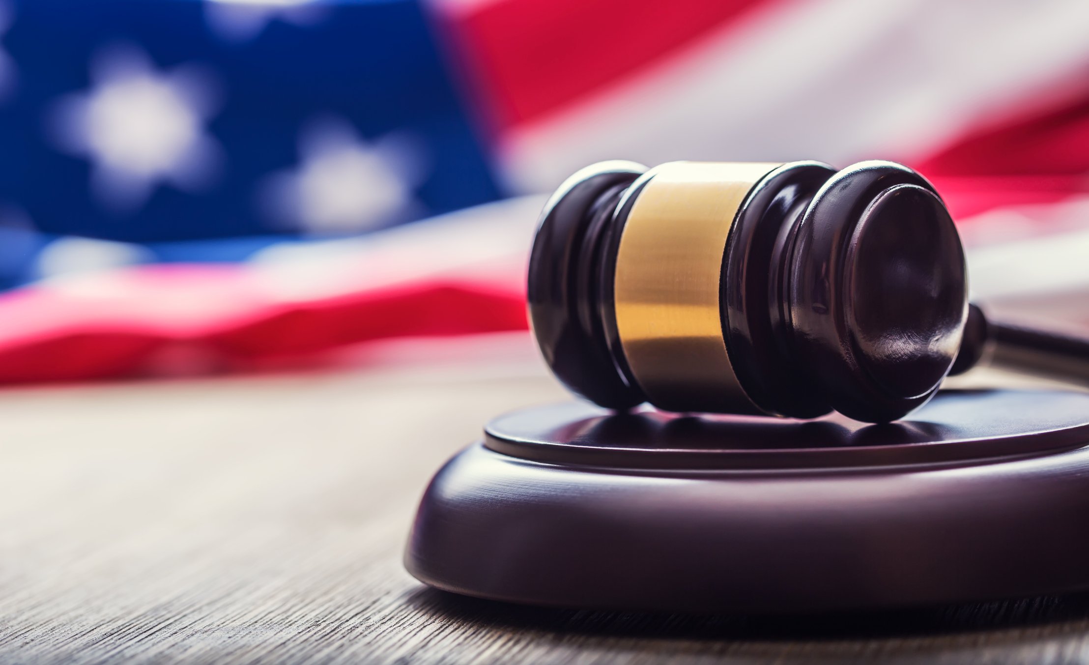 Close-up photo of a gavel resting on a wooden stand in front of an out-of-focus American flag.