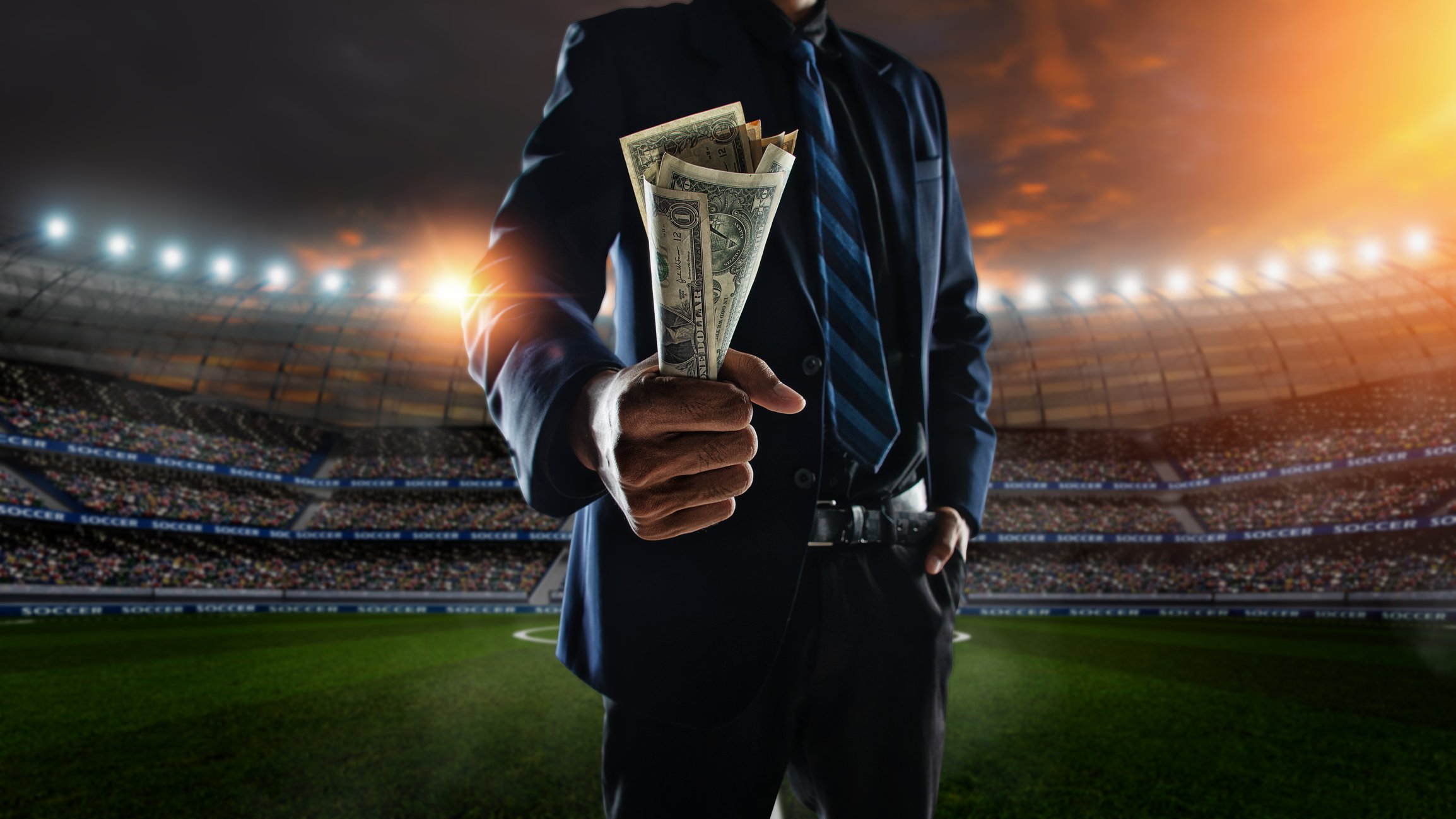 a man in a suit holds up a wad of money while standing in front of a shadowed image of a sports stadium full of people